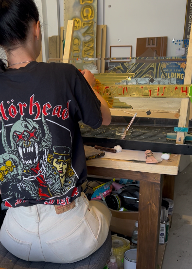 A person working at a table in a workshop, wearing a black t-shirt with a graphic design related to heavy metal, creating or assembling a decorative piece.