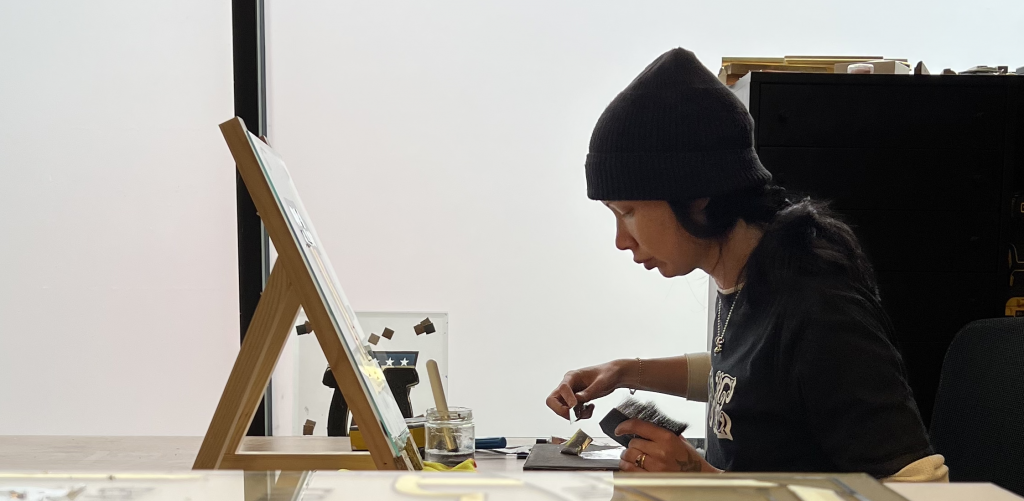 A person working diligently on a gilded mirror at an easel in a workshop, surrounded by tools and materials.